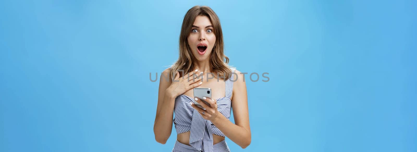 Woman feeling surprised and grateful after reading unexpected news from smartphone pressing palm to chest from amazement dropping jaw and smiling staring at camera, cannot believe it happened. Technology concept