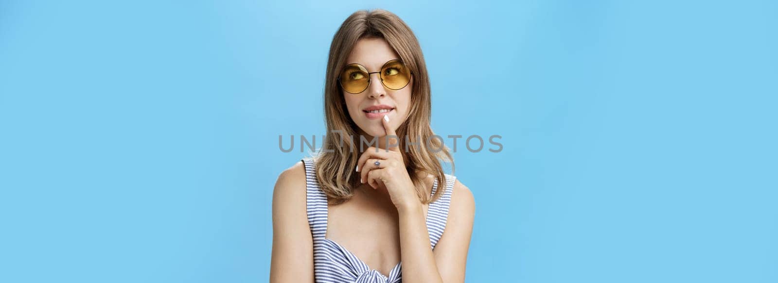 Flirty and creative charming feminine woman in stylish sunglasses and top holding finger on lip gazing at upper right corner thoughtful making up plan or thinking about tasty cake she wants by Benzoix