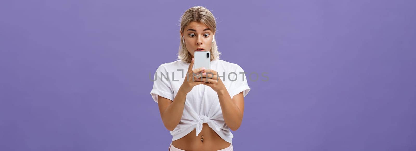 Girl watching thrilling video on social media in wireless earphones holding smartphone near face popping eyes at smartphone screen gazing at gadget with interest and curiosity over purple background by Benzoix