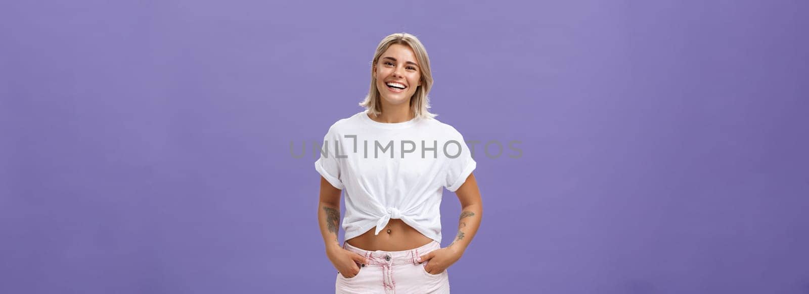 Joyful attractive tanned girlfriend with blond hair and tattoos on arms with cropped top chilling by pool with friends talking with amused happy expression smiling at camera holding hands in pockets by Benzoix