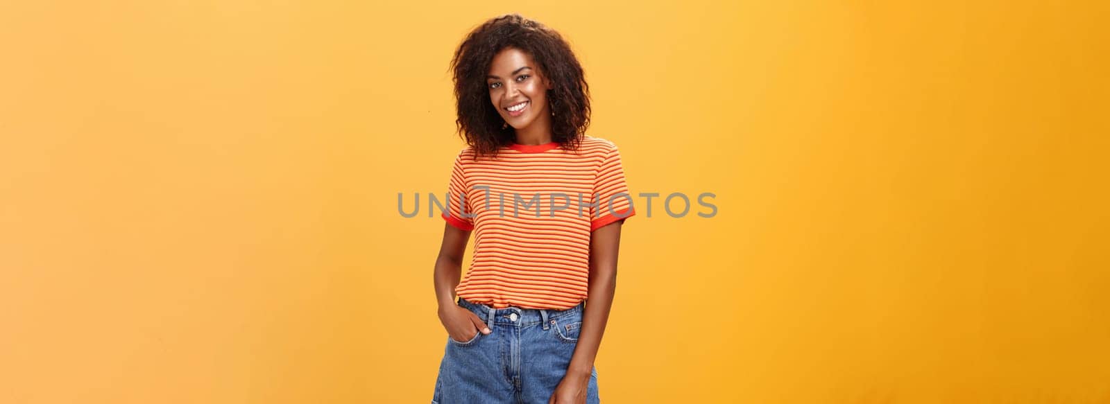 Ready to travel world. Energetic confident and attractive dark-skinned woman with curly hair holding hand in pocket of denim shorts smiling joyfully posing over orange background carefree and friendly.