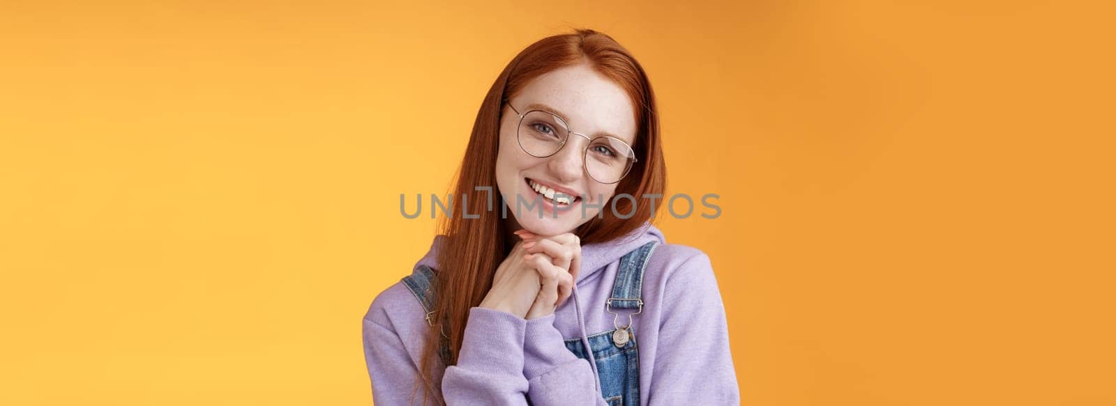 Tender romantic lovely redhead girlfriend leaning palms tilting head cute flirty giggling smiling camera white teeth wearing glasses hoodie overalls near orange background, sensually laughing.