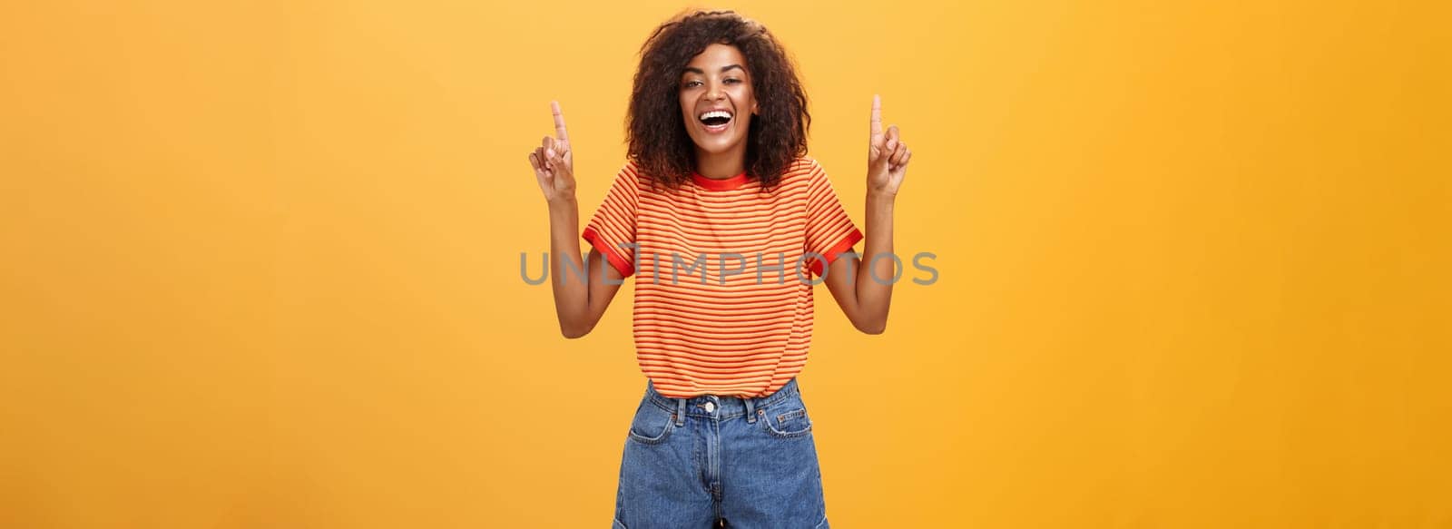 Woman feeling amused and entertained. Portrait of happy carefree stylish African-American girl with afro hairstyle laughing out loud joyfully pointing up with raised arms over orange wall by Benzoix
