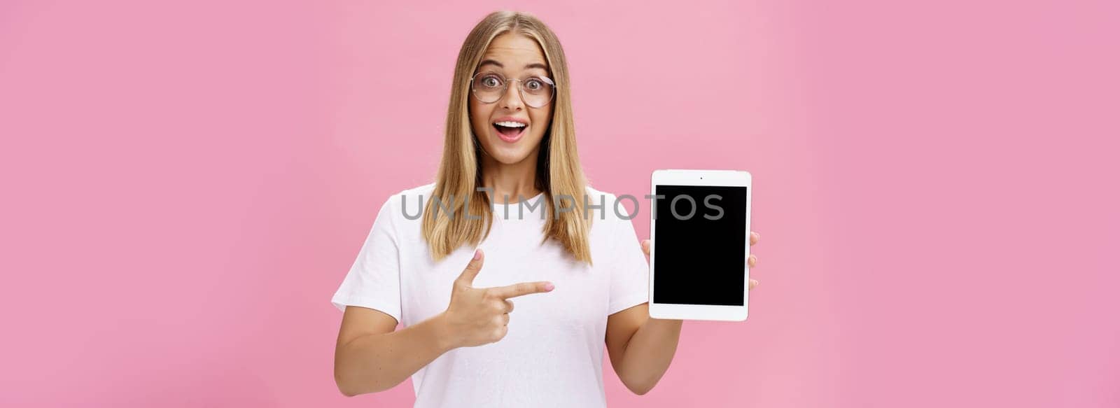 Girl suggests buy digital tablet for university and forget paper books. Excited happy and delighted young female student in white t-shirt and glasses pointing at device screen recommending gadget by Benzoix