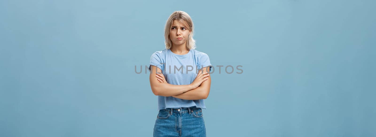 Uncertain troubled and perplexed attractive blond woman with tanned skin holding hands crossed on chest pouting and frowning looking right with worried unsure and doubtful look over blue wall by Benzoix