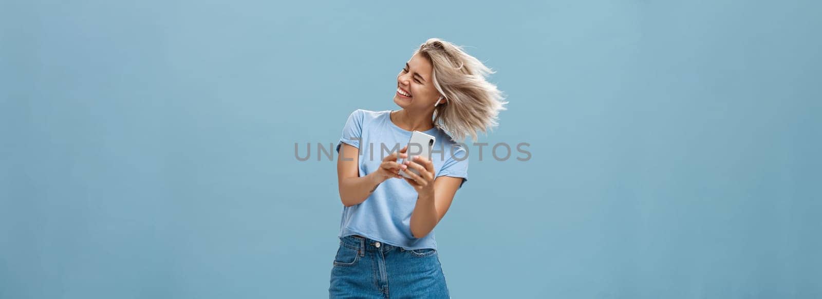 Great vibes carrying me away. Carefree joyful attractive young blonde female in stylish outfit waving head and smiling broadly listening music in wireless earbuds and holding smartphone over blue wall. Emotions and technology concept