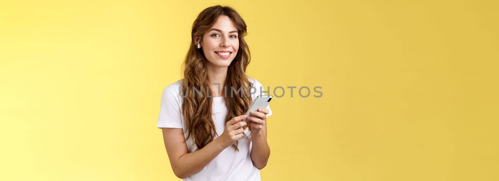 Feminine lovely curly-haired woman white t-shirt hold smartphone picked awesome new song listen music wearing wireless earphones smiling delighted camera enjoy earbuds beats yellow background by Benzoix