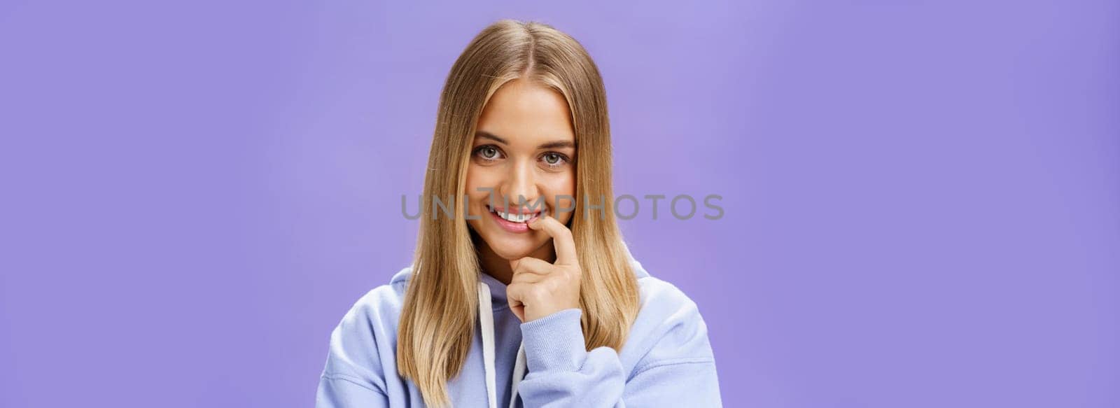 Close-up shot of feminine and tender young girl with fair hair and tanned skin biting finger flirty and shy smiling cute at camera standing in trendy over-sized hoodie, flirting over purple background by Benzoix