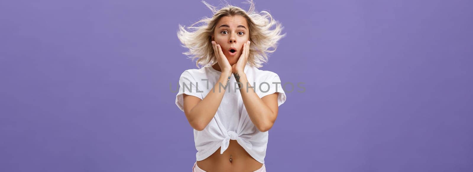 Romantic surprised and thrilled feminine blonde female with tattoos standing on windy street folding lips and holding hands on face impressed and intrigued while hair floats in air over purple wall by Benzoix