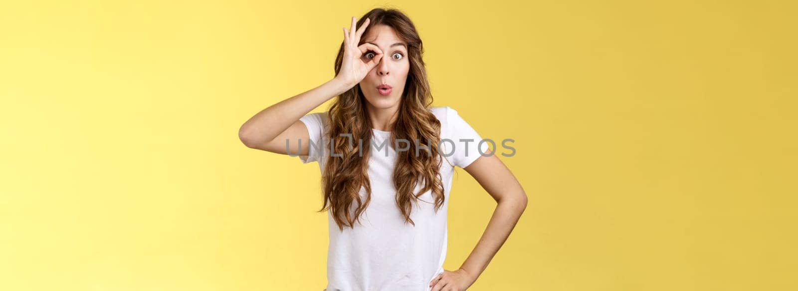 Attractive young astonished curly-haired european girl fascinated folding lips admiration stare alluring thing spot awesome promo hold ring gesture okay ok through eye check out interesting spot. Lifestyle.