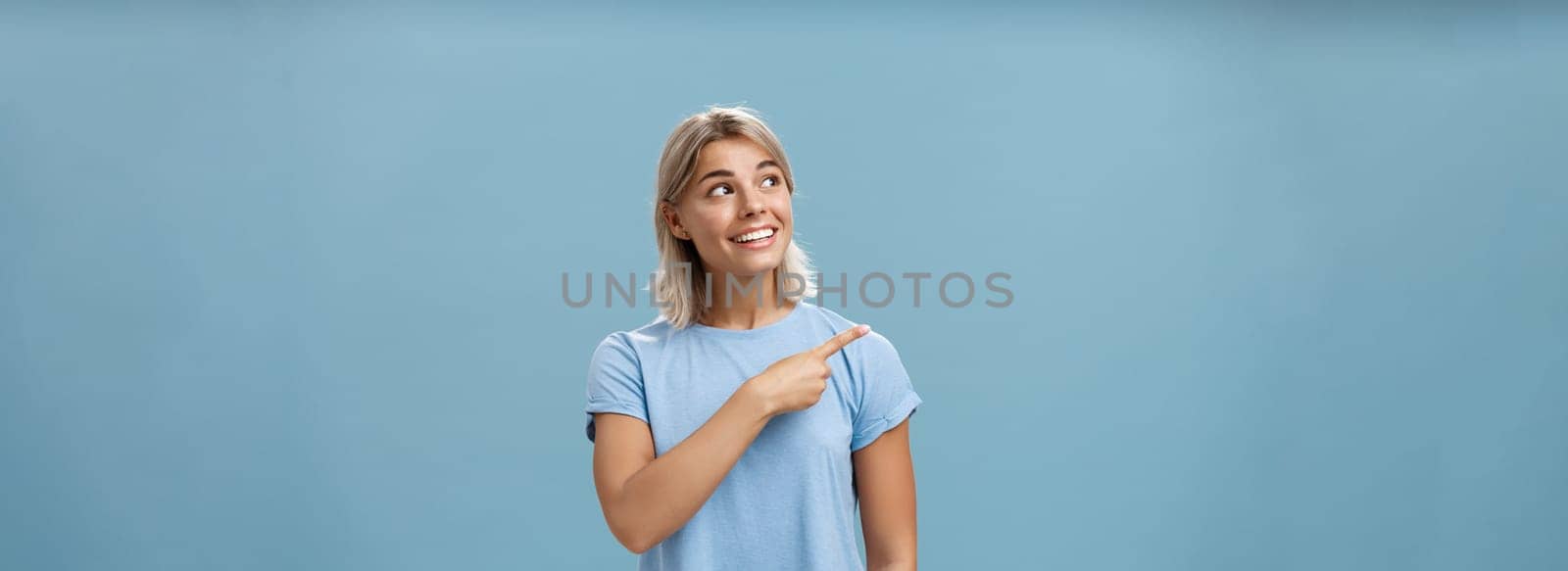 Lifestyle. Waist-up shot of creative and dreamy happy attractive blonde female in casual t-shirt looking and pointing at upper left corner with admiration and joy standing over blue background amused and curious.