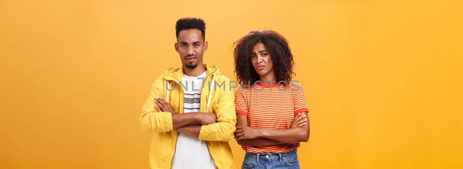Two friends dislike lame idea of mate. Portrait of dissatisfied unimpressed african american man and woman crossing arms on chest in aversion frowning doubtful and disappointed over orange wall.