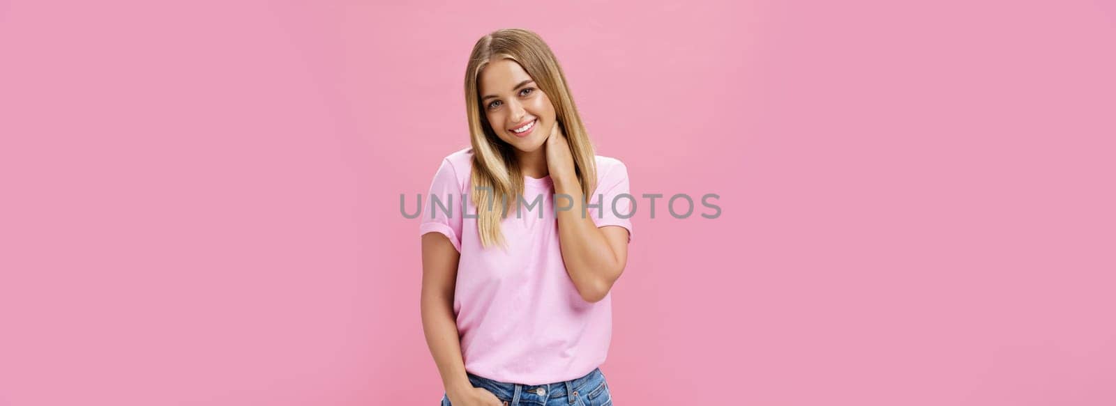 Portrait of shy and timid attractive european woman with fair hair in casual outfit touching neck silly tilting head and smiling cute at camera standing over pink background. Emotions concept