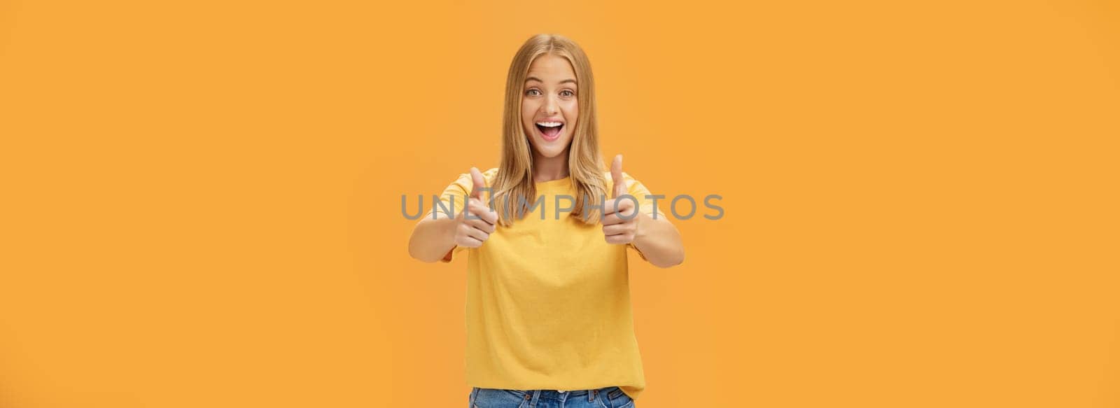 Woman supports with raised thumbs up and amused cheerful smile showing positive attitude expressing like on concept or idea giving approval posing happy and delighted against orange background. Lifestyle.