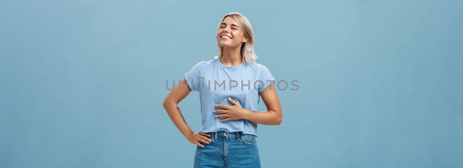 Cute girl likes eat after workout being pleased and stuffed after dinner. Portrait of satisfied attractive blonde young woman touching belly raising head up with broad smile over blue background by Benzoix