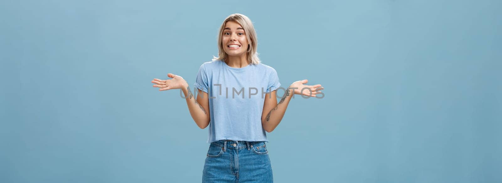 Oops I do not know. Silly cute european girl with fair hair and cool tattoos shrugging with palms spread aside in no idea gesture smiling with sorry look being unaware and clueless over blue wall by Benzoix