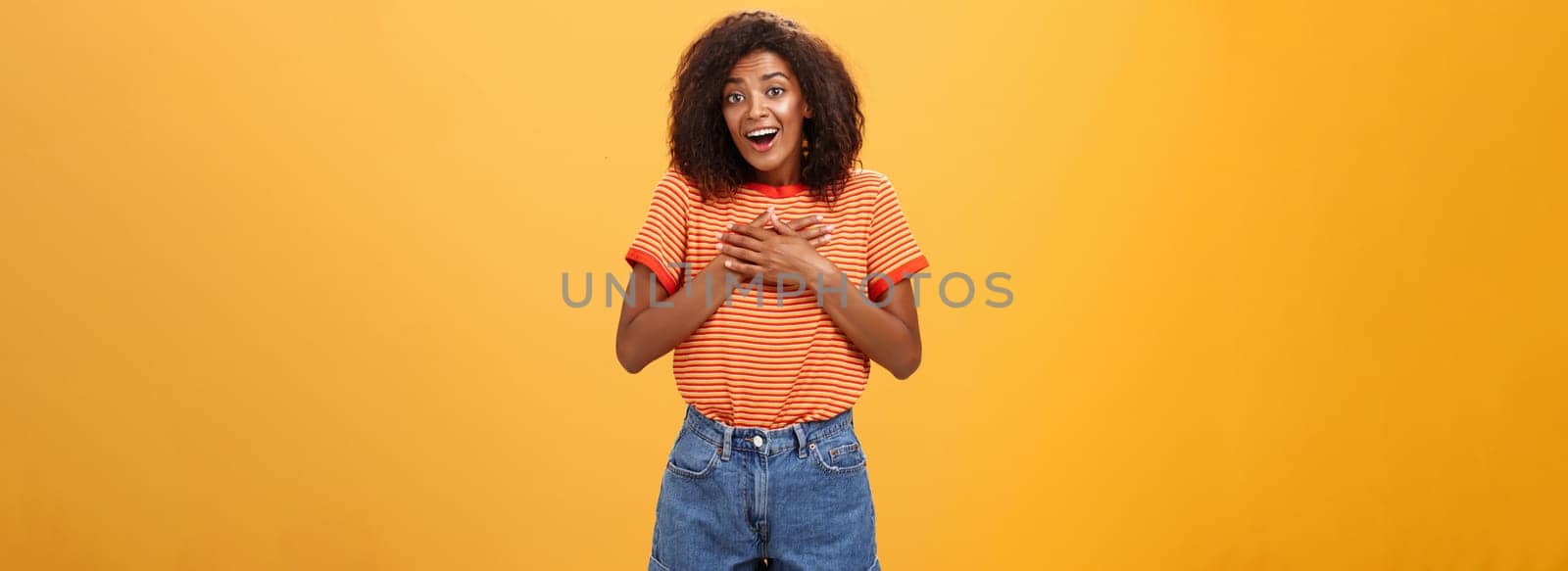 Cute delighted african american woman being grateful and thankful for help standing pleased over orange wall holding palms on breast thanking friend for rescuing grinning joyfully at camera by Benzoix