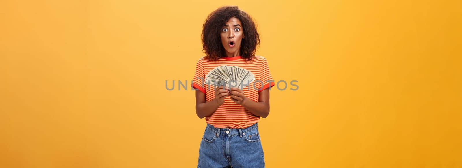Woman shocked finding lots of cash in safe. Portrait of surprised speechless good-looking dark-skinned female with curly haircut folding lips gasping holding money posing over orange background.