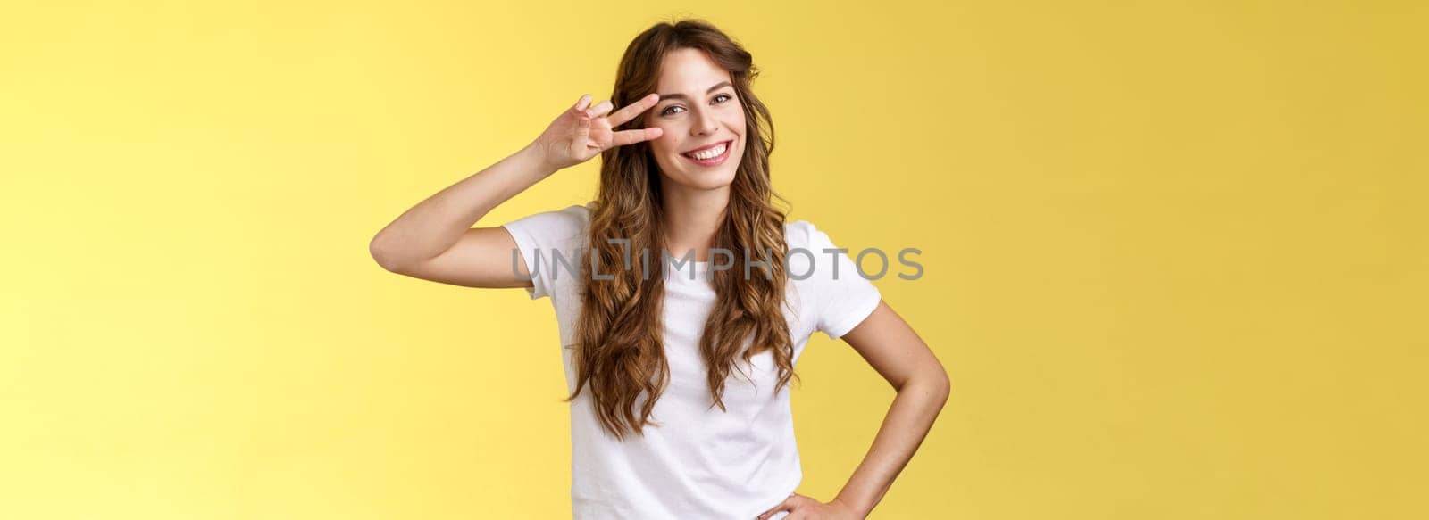 Positive outgoing enthusiastic good-looking woman having fun enjoy weekend summer vacation travel show peace sign victory gesture on eye tilt head having fun smiling broadly yellow background. Lifestyle.