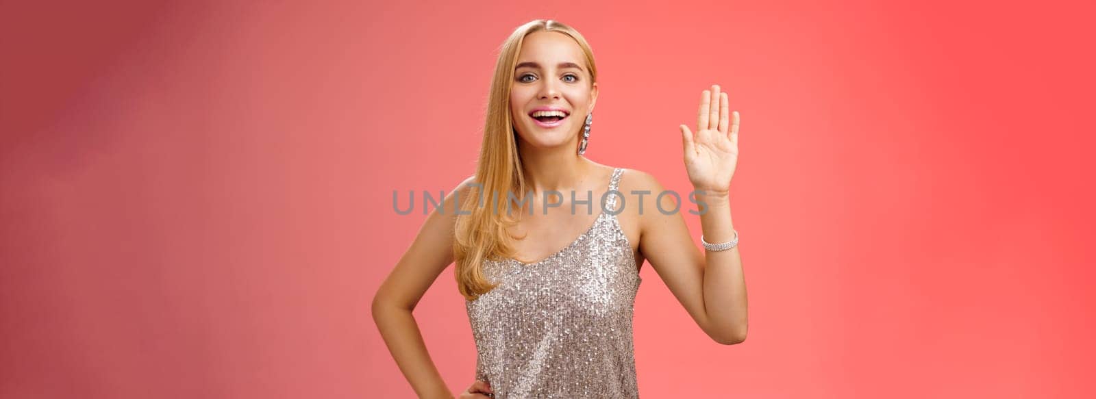 Lifestyle. Friendly stylish glamour attractive blond caucasian woman in silver glittering dress hold hand waist waiving palm hello hi what up gesture smiling introducing herself party boyfriend friend.