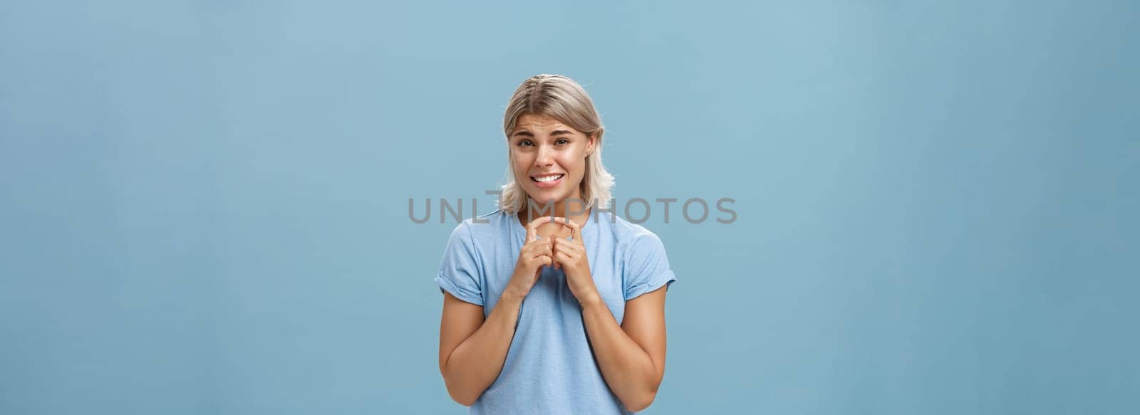 Silly cute and worried blonde female friend with short haircut steepling index fingers and smiling with guilty sorry smile frowning while waiting for answer begging for help or favor over blue wall by Benzoix