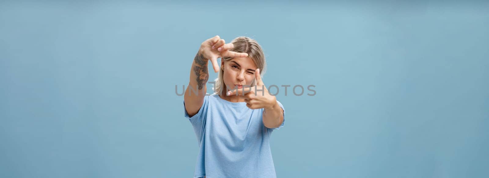 Artistic and creative female designer in stylish t-shirt closing one eye folding lip and making frame gesture while looking through it as if taking measurement or picturing something over blue wall. Copy space