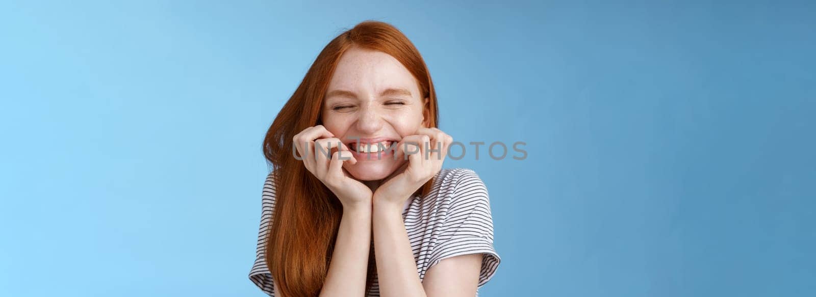 Cheerful carefree giggling ginger girl look happy bright close eyes smiling delighted hold hands cheeks having fun feeling excitement joy triumphing cheering good news, standing blue background.