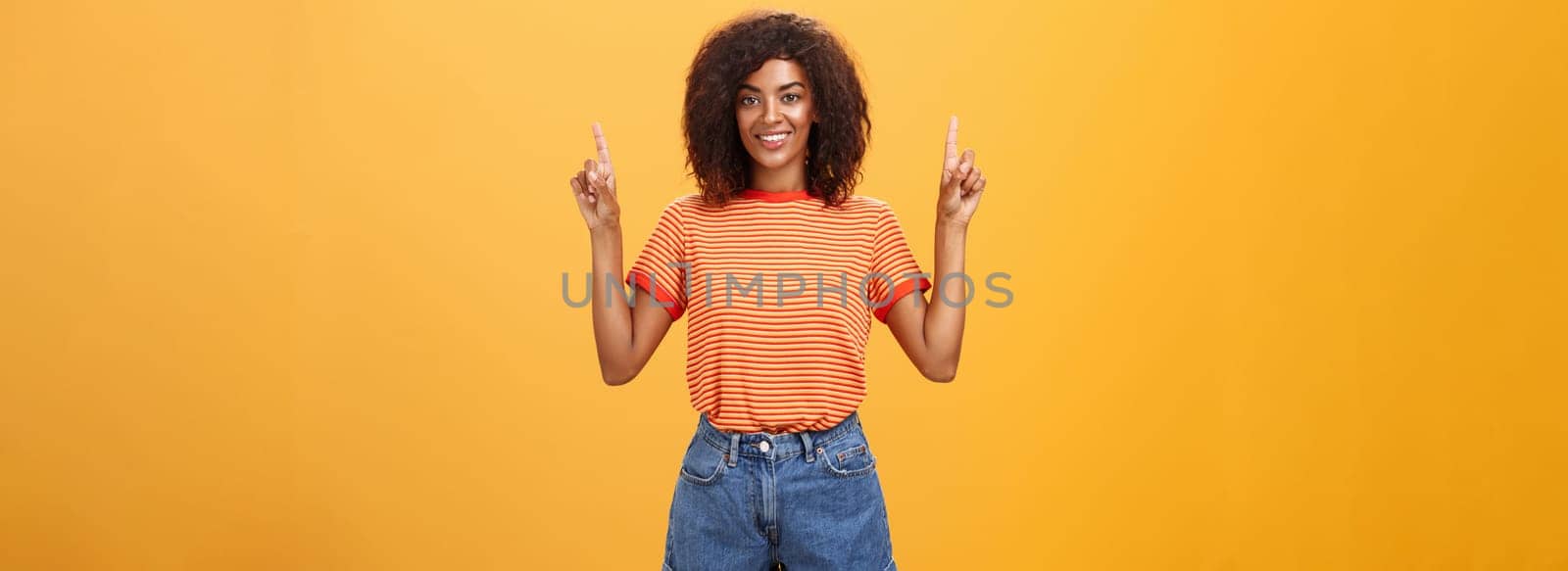 Looking only up and forward. Optimistic ambitious stylish dark-skinned female student in striped cool t-shirt and shorts raising hands pointing upwards and smiling friendly over orange wall.