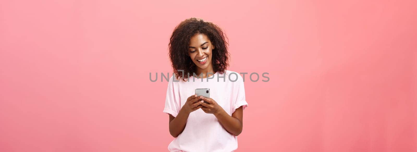 Girl writing text message to friend. Portrait of charming amused and happy young dark-skinned woman with curly hair looking at smartphone screen typing smiling enjoying cool features of cellphone.