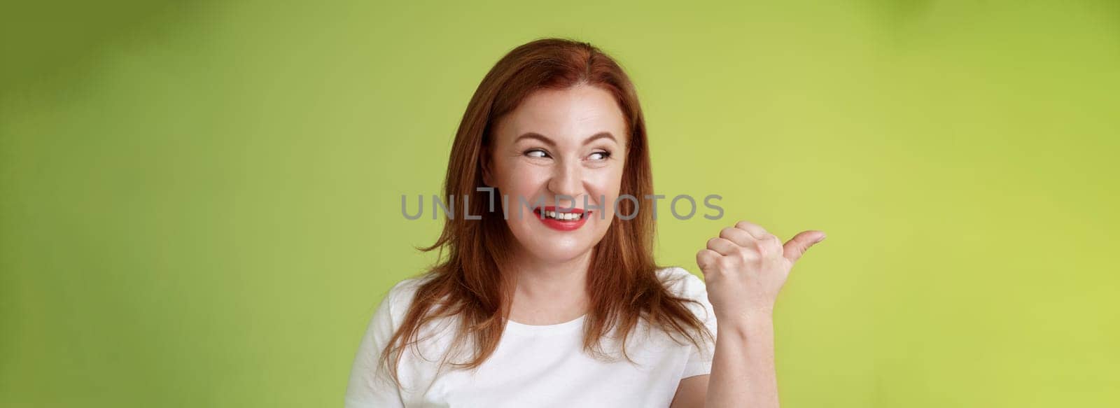 Intrigued charismatic redhead middle-aged woman smiling temtation interest pointing looking left curiously check-out cool promo share awesome place location stand green background by Benzoix
