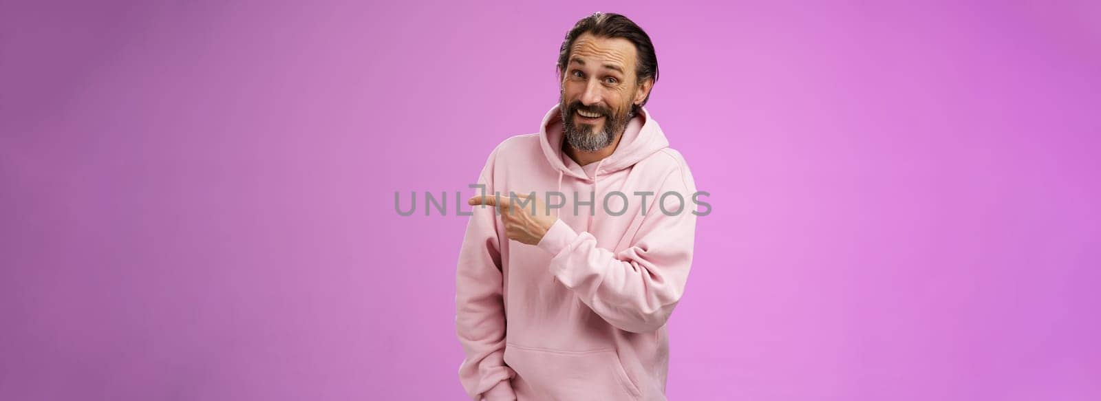 Friendly carefree caucasian bearded man in pink hoodie pointing right intoducing family members smiling proudly delighted showing blank copy space advertising, posing purple background by Benzoix