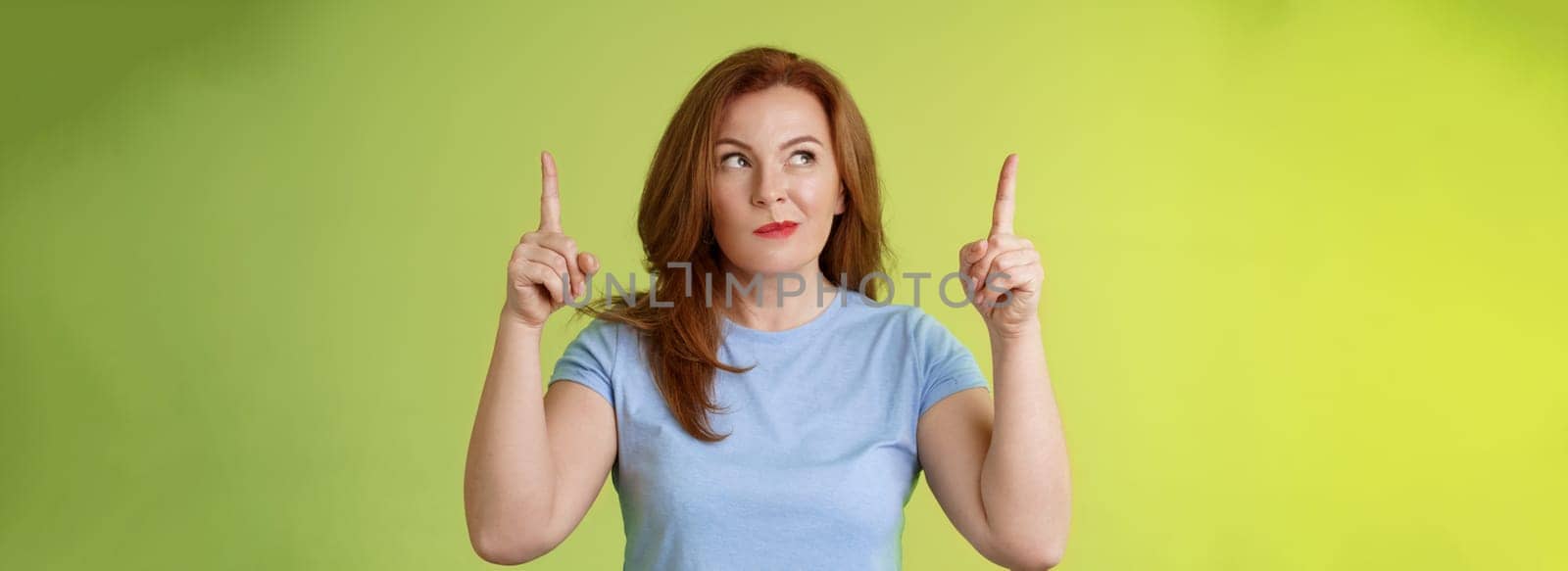 Curious thoughtful cute redhead female middle-aged ginger mother smirking thinking look aside pointing up index fingers deciding choice pondering choosing between variants green background by Benzoix