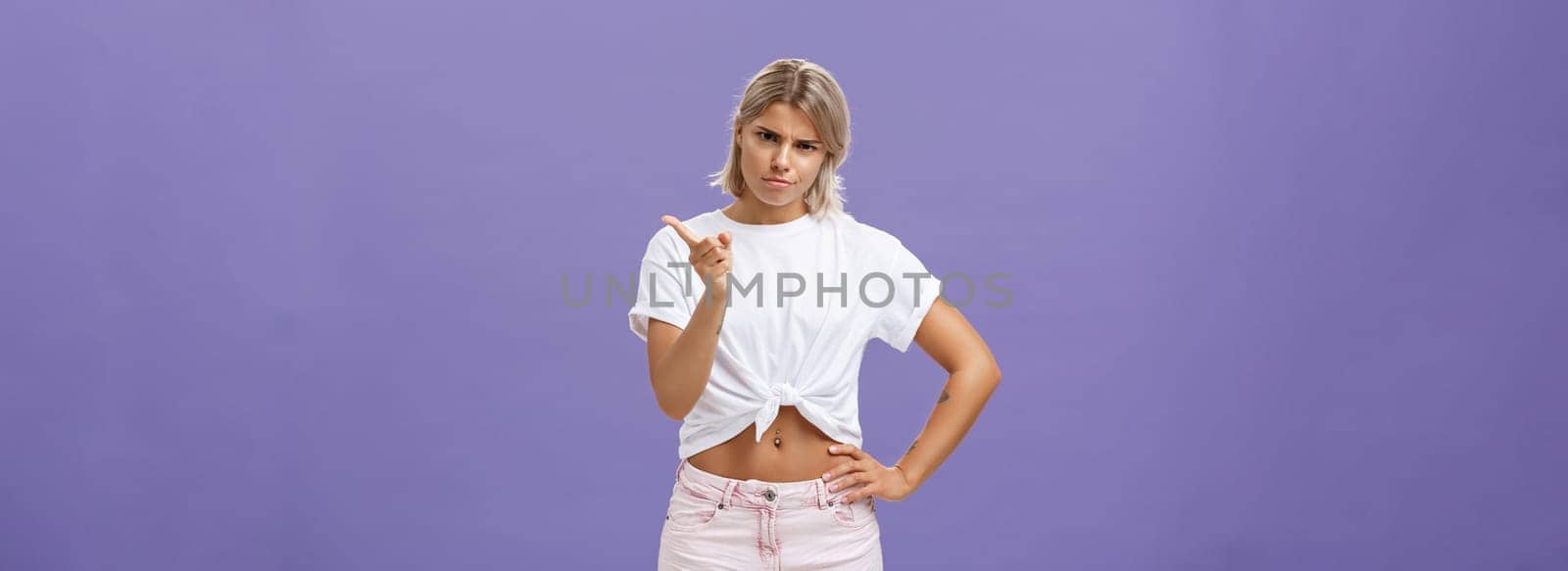 I told you behave properly. Strict and cute displeased blonde female in white t-shirt and shorts shaking index finger in strict pose looking from under forehead while scolding over blue background.