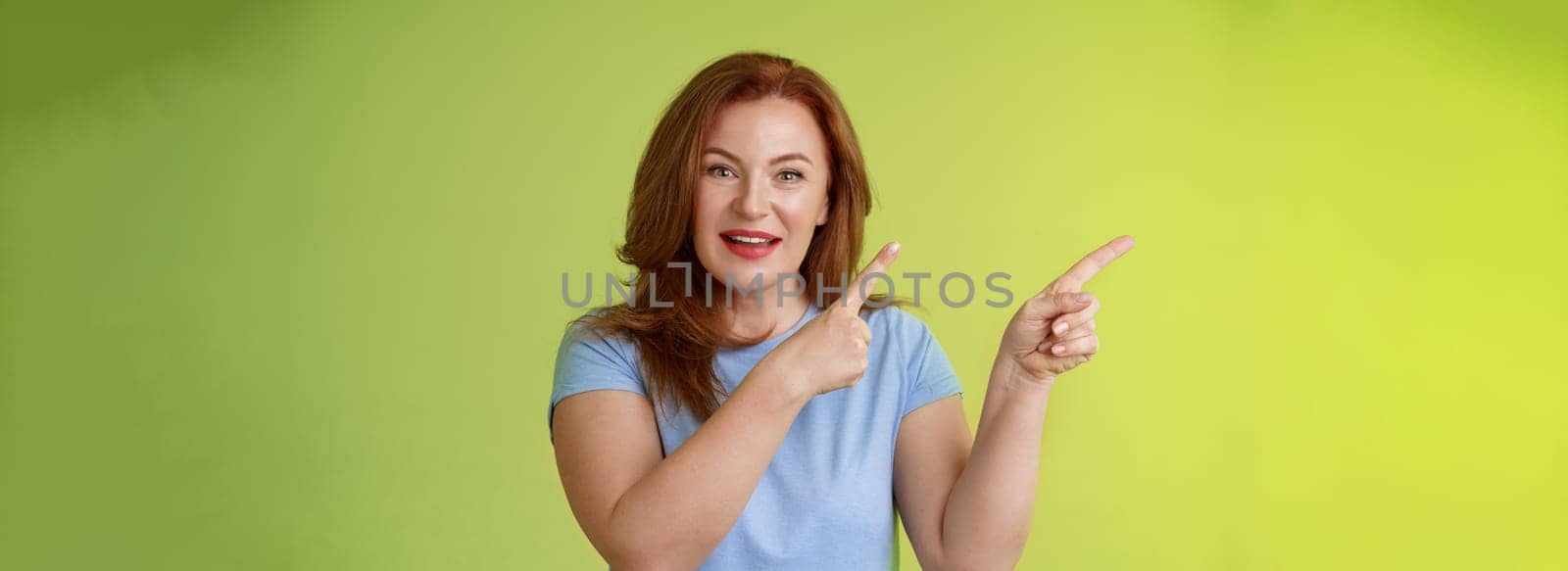 Curious enthusiastic gorgeous redhead female intrigued pointing upper left corner asking question interesting product talk shop assistant consulting promo stand green background blue t-shirt.