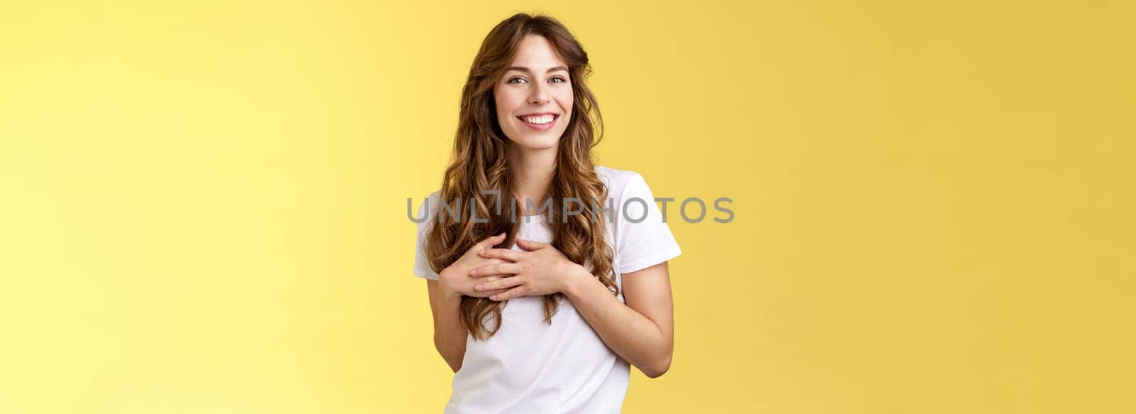 Lovely joyful tender feminine girlfriend long curly hairstyle touch heart feel heartbeat delighted receive lovely heartwarming gift smiling broadly satisfied flirty gazing camera yellow background.
