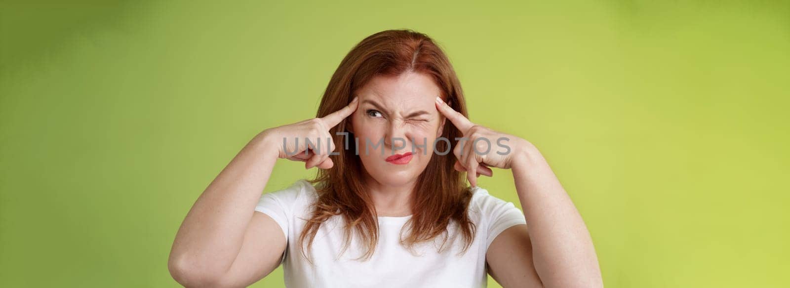 Difficult decision. Perplexed hesitant redhead middle-aged 50s woman trying solve puzzle look puzzled troubled smirk close eyes peek right touch temples thoughtful thinking intense green background.