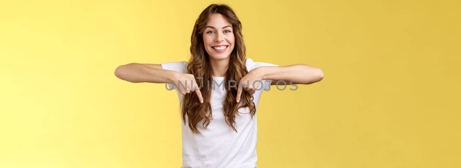 Friendly cheerful excited happy smiling girl long natural brunette haircut grinning joyfully pointing down index fingers show friend great choice introduce promo advice click link yellow background. Lifestyle.