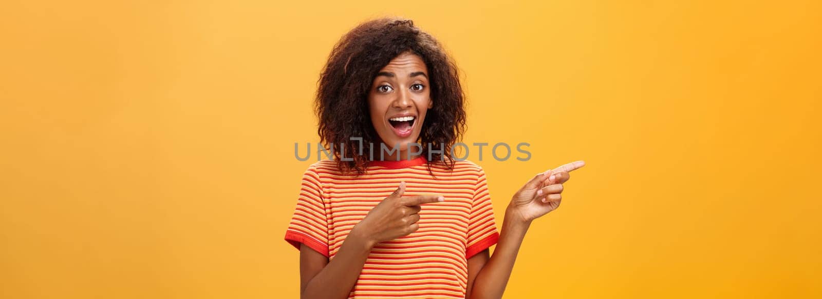 Portrait of amazed excited charismatic dark-skinned young pretty girl with afro hairstyle in trendy striped t-shirt pointing left delighted and fascinated posing against orange background. Lifestyle.