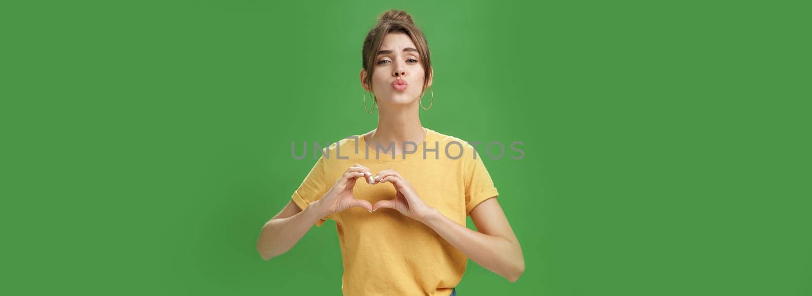 Sensual charming woman in yellow t-shirt and combed hairstyle in yellow t-shirt showing heart gesture against chest folding lips to give kiss or mwah tender and loving over green background. Lifestyle.