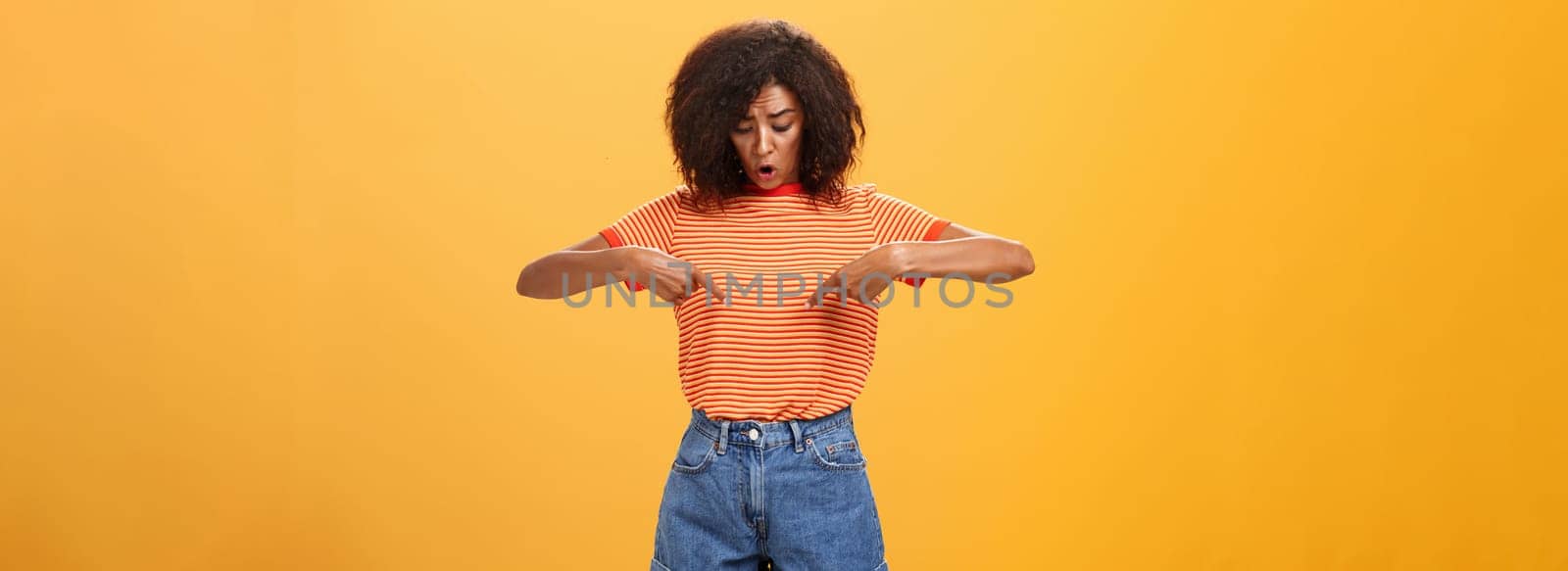 Portrait of worried questioned and surprised african american female with curly hairstyle looking and pointing at stomach or belly feeling discomfort and problems with health over orange background. Lifestyle.