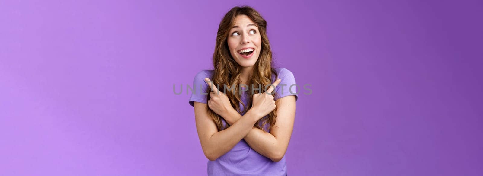 Happy cheerful lively curly girl smiling broadly receive awesome opportunity study abroad choosing universities cross hands sideways pointing left right grinning amused entertained purple wall.