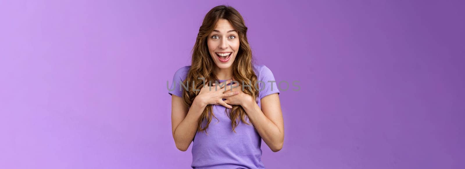 Surprised happy pleased cheerful girl brown curly hairstyle press hands heart amused delighted thanking friend adorable lovely gift smiling broadly grateful gaze camera stand purple background. Lifestyle.