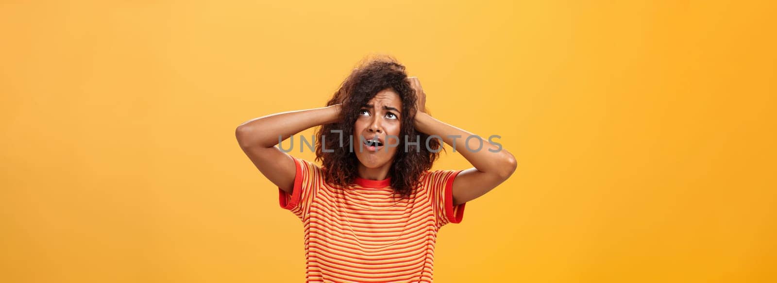 Displeased moody african girlfriend holding hands on head and frowning looking concerned and dissatisfied up in sky panicking rain will spoil hairstyle standing nervous over orange background by Benzoix