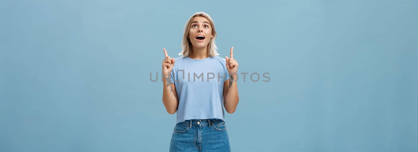 Indoor shot of impressed speechless attractive fair-haired female student in casual t-shirt and denim shorts dropping jaw from amazement pointing and looking up intrigued over blue wall by Benzoix