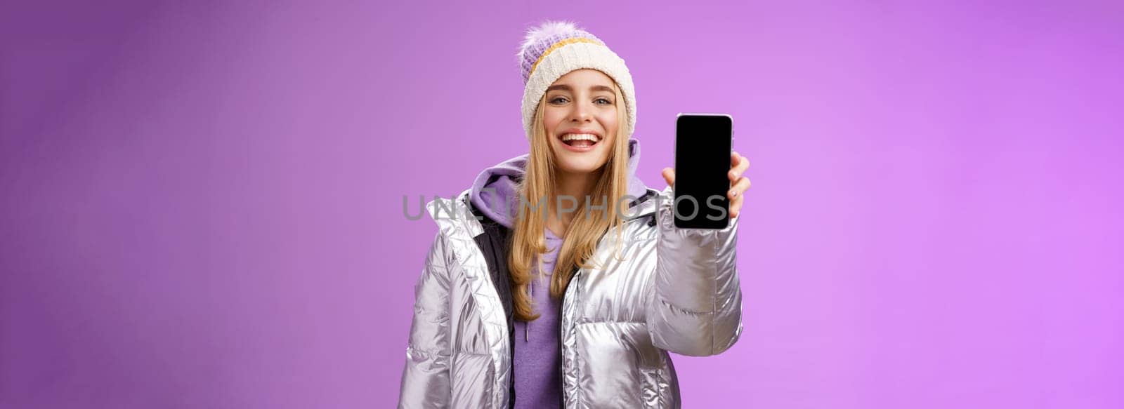 Sassy delighted cheerful blond woman recommend use app edit perfect pictures holding smartphone showing mobile phone display proudly satisfactory smile camera, standing purple background.