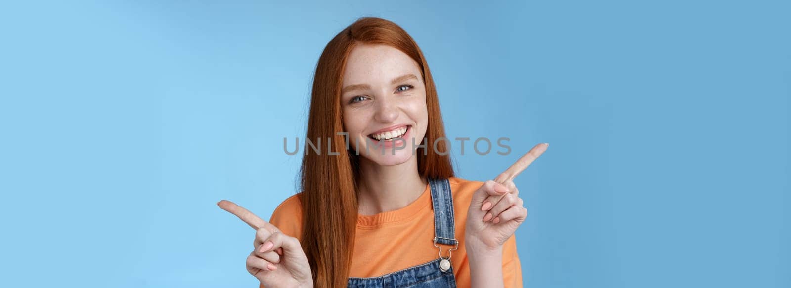 Friendly happy laughing pretty redhead girl female student showing lots opportunities advice make choice pointing sideways left right introducing different product grinning gladly, blue background.