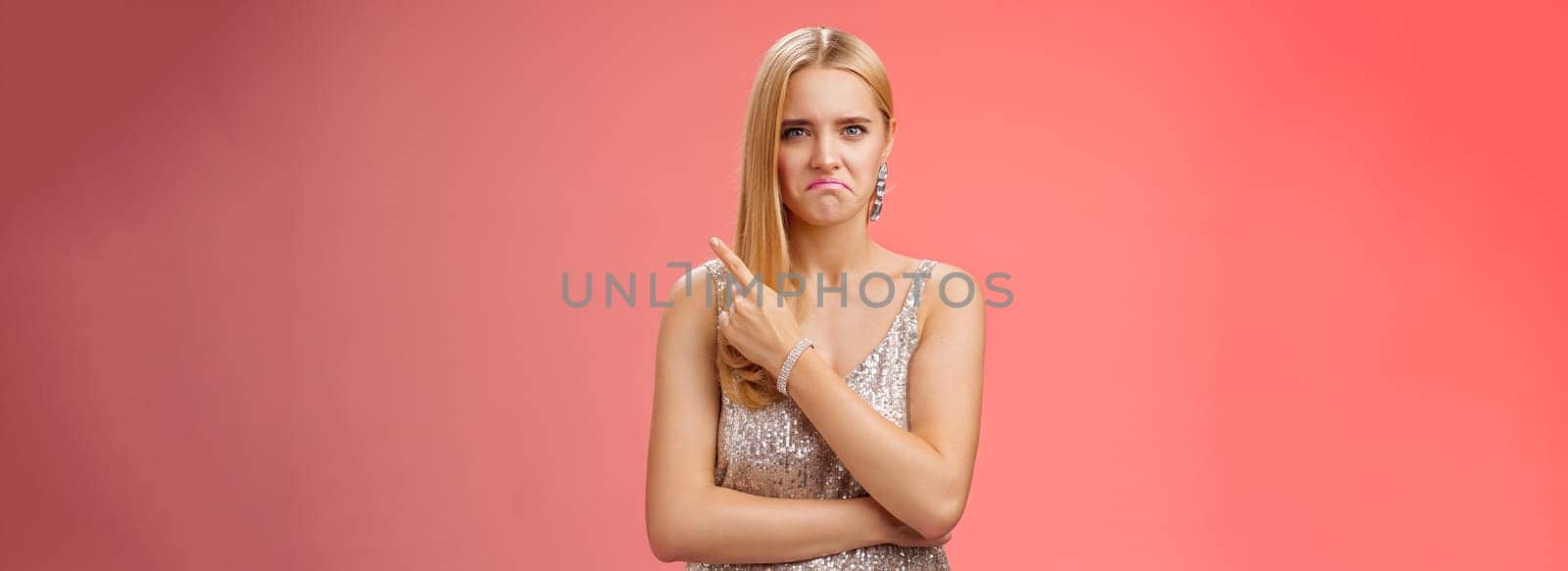 Lifestyle. Jealous displeased angry young revengeful blond ex-girlfriend in luxurious silver shiny dress frowning pouting pointing upper right corner displeased pissed standing fed up upset red background.