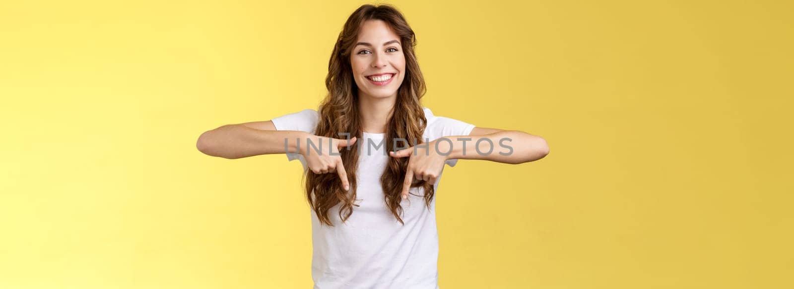 Lively helpful good-looking caucasian girl curly long hair showing you excellent place find hairdresser pointing down index fingers smiling toothy happy delighted grin stand yellow background by Benzoix