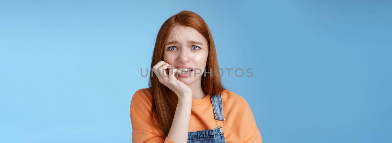 Intense worried scared unsure young redhead panicking silly girl frowning look upset anxious biting fingernails emotional frightened fired standing blue background emotional terrified by Benzoix