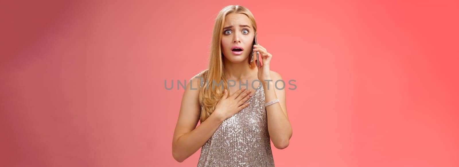 Concerned worried woman blond hairstyle in silver stylish glittering dress hold hand heart widen eyes afraid gasping speechless listen terrifying news talking smartphone express empathy worry by Benzoix